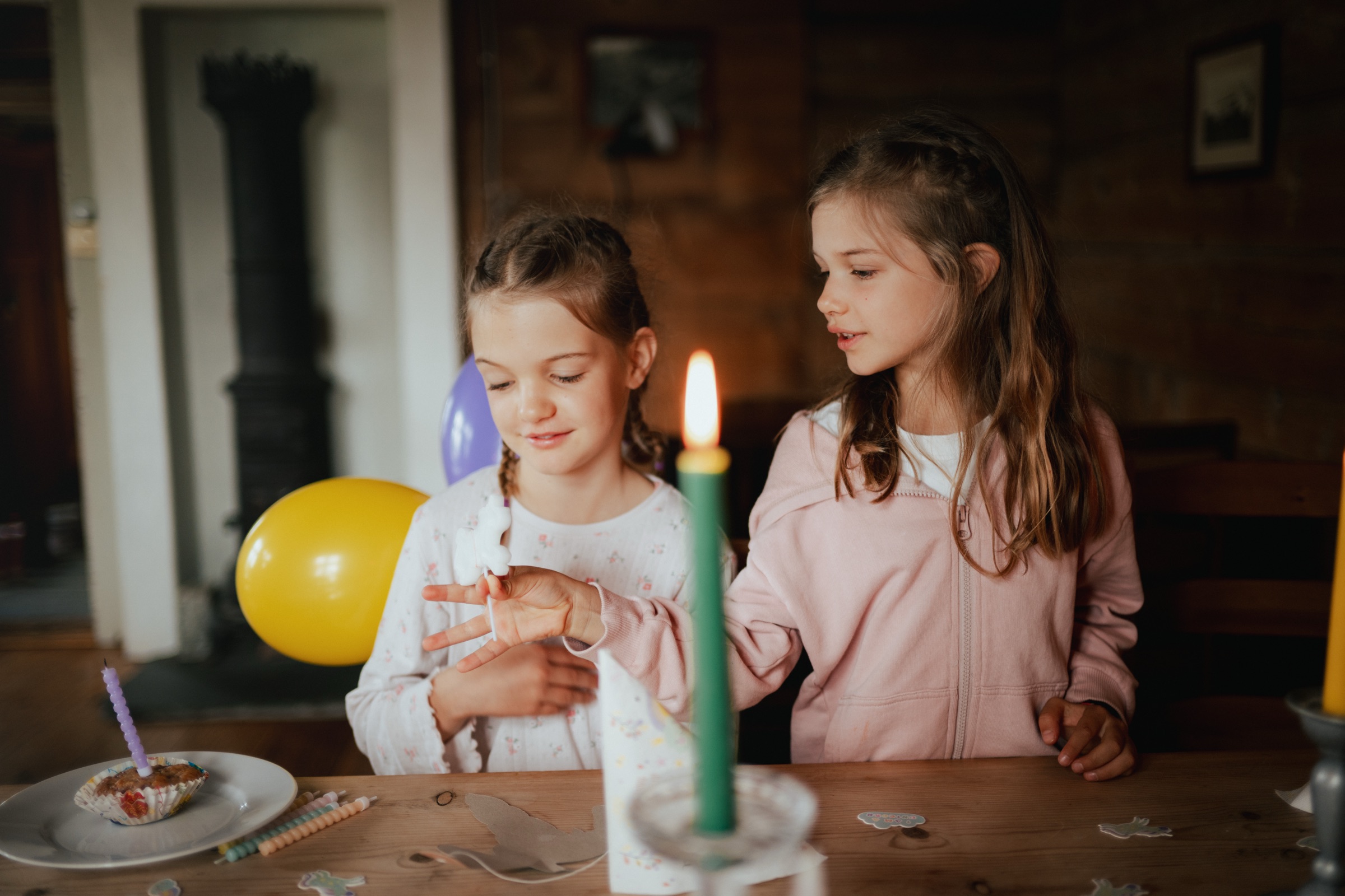 Zwei Kinder sitzen im Kerzenlicht am Tisch, eines hält einen gelben Luftballon.